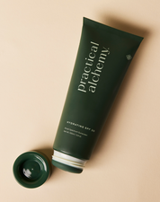 Open green tube of Practical Alchemy Hydrating SPF 30 sunscreen with lid next to it on a beige background