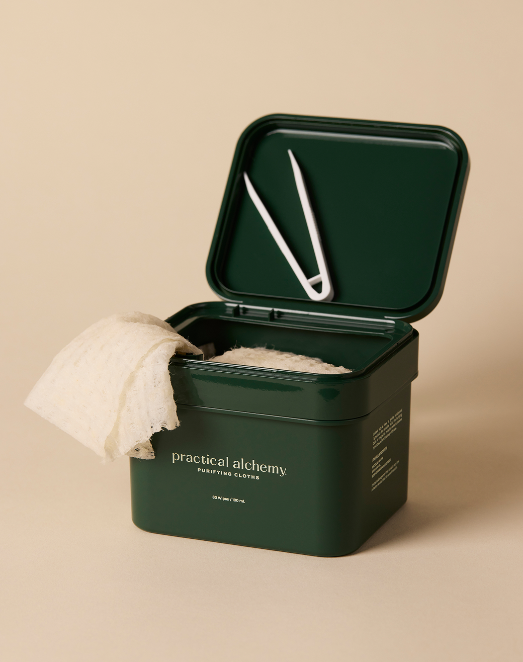 Green container with a white purifying cloths and a white tweezers on a beige background, featuring 'practical alchemy' branding.