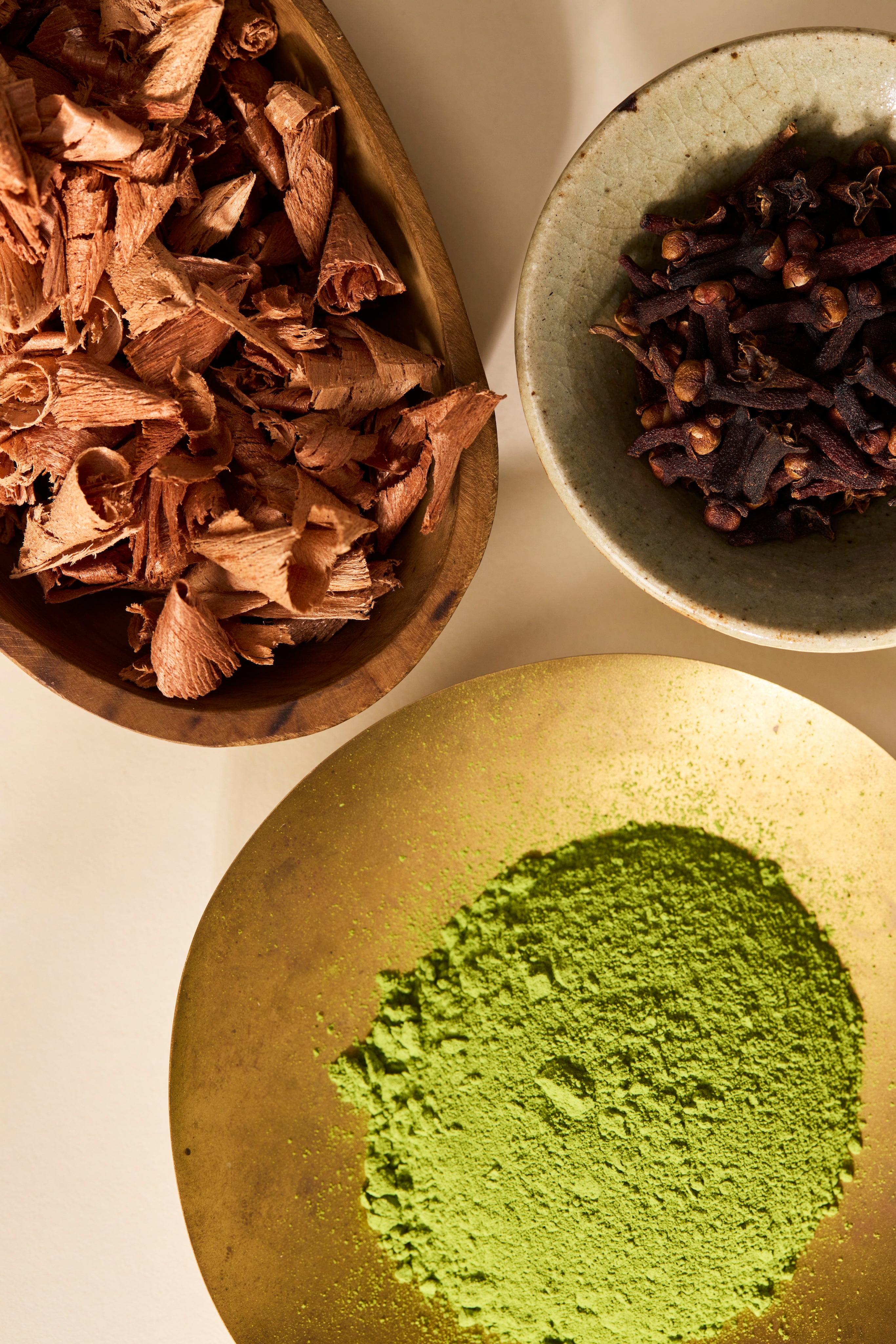 Three bowls containing sandlewood, dried cloves, and green matcha powder on a light background.